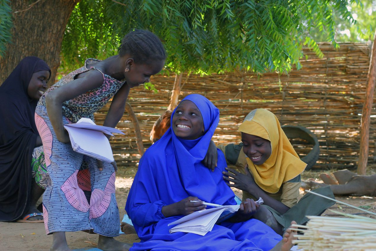 Tre piger laver lektier i skyggen af et tre. De smiler til hinanden.